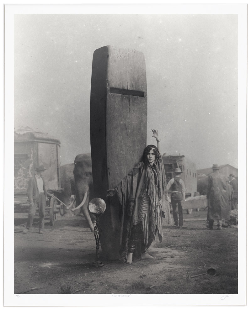 Black-and-white surreal photograph of a woman posed beside a tall monolithic structure, with elephants and workers in the background.